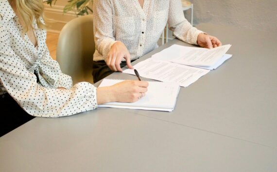 woman signing on white printer paper beside woman about to touch the documents